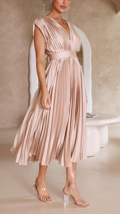 Champagne - A woman wearing a dusty rose pleated satin midi dress with a deep V-neckline, cap sleeves, and a cinched waist, standing against a textured stone wall.