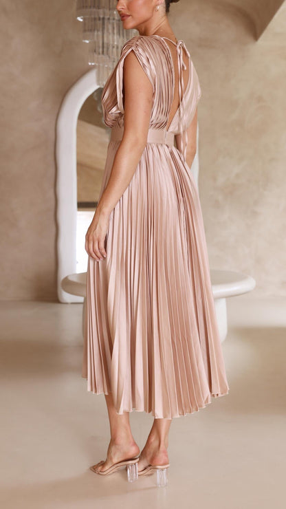 Champagne - A woman wearing a dusty rose pleated satin midi dress with a deep V-neckline, cap sleeves, and a cinched waist, standing against a textured stone wall.