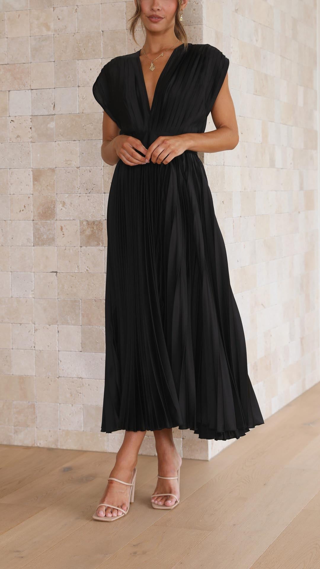 Champagne - A woman wearing a dusty rose pleated satin midi dress with a deep V-neckline, cap sleeves, and a cinched waist, standing against a textured stone wall.