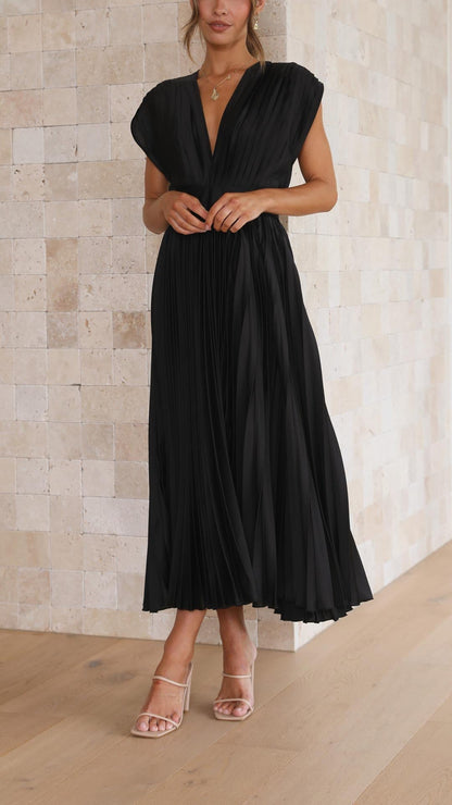 Champagne - A woman wearing a dusty rose pleated satin midi dress with a deep V-neckline, cap sleeves, and a cinched waist, standing against a textured stone wall.