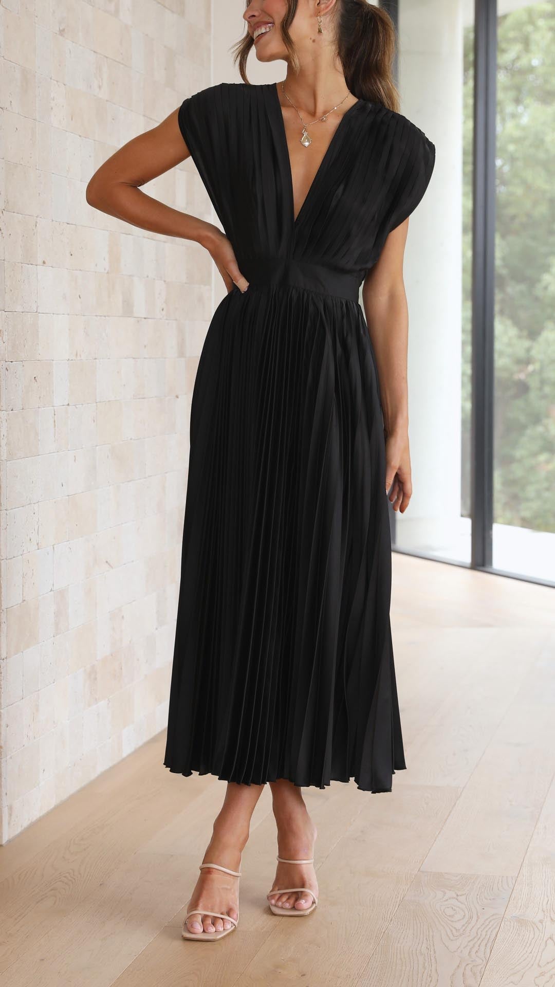 Champagne - A woman wearing a dusty rose pleated satin midi dress with a deep V-neckline, cap sleeves, and a cinched waist, standing against a textured stone wall.