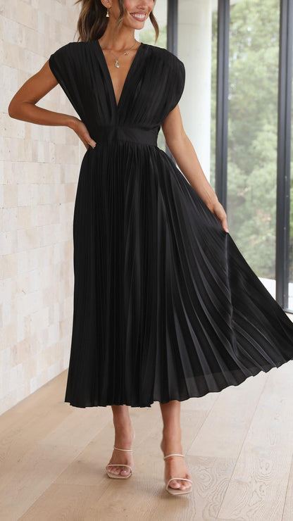 Champagne - A woman wearing a dusty rose pleated satin midi dress with a deep V-neckline, cap sleeves, and a cinched waist, standing against a textured stone wall.