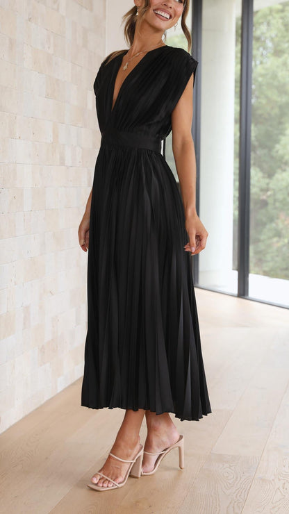 Champagne - A woman wearing a dusty rose pleated satin midi dress with a deep V-neckline, cap sleeves, and a cinched waist, standing against a textured stone wall.