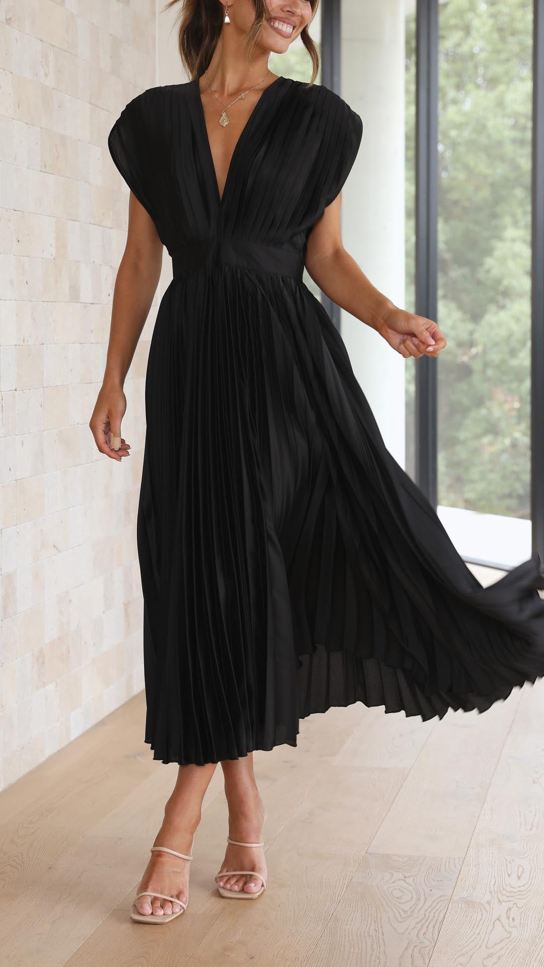 Black - A woman wearing a dusty rose pleated satin midi dress with a deep V-neckline, cap sleeves, and a cinched waist, standing against a textured stone wall.