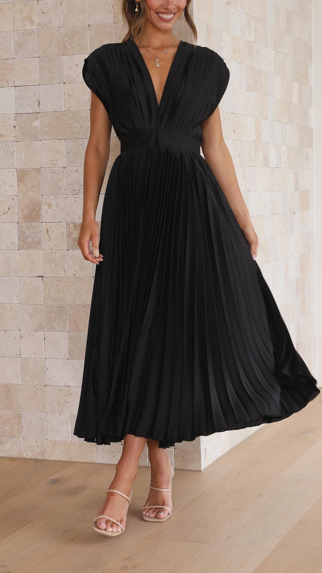 Black - A woman wearing a dusty rose pleated satin midi dress with a deep V-neckline, cap sleeves, and a cinched waist, standing against a textured stone wall.