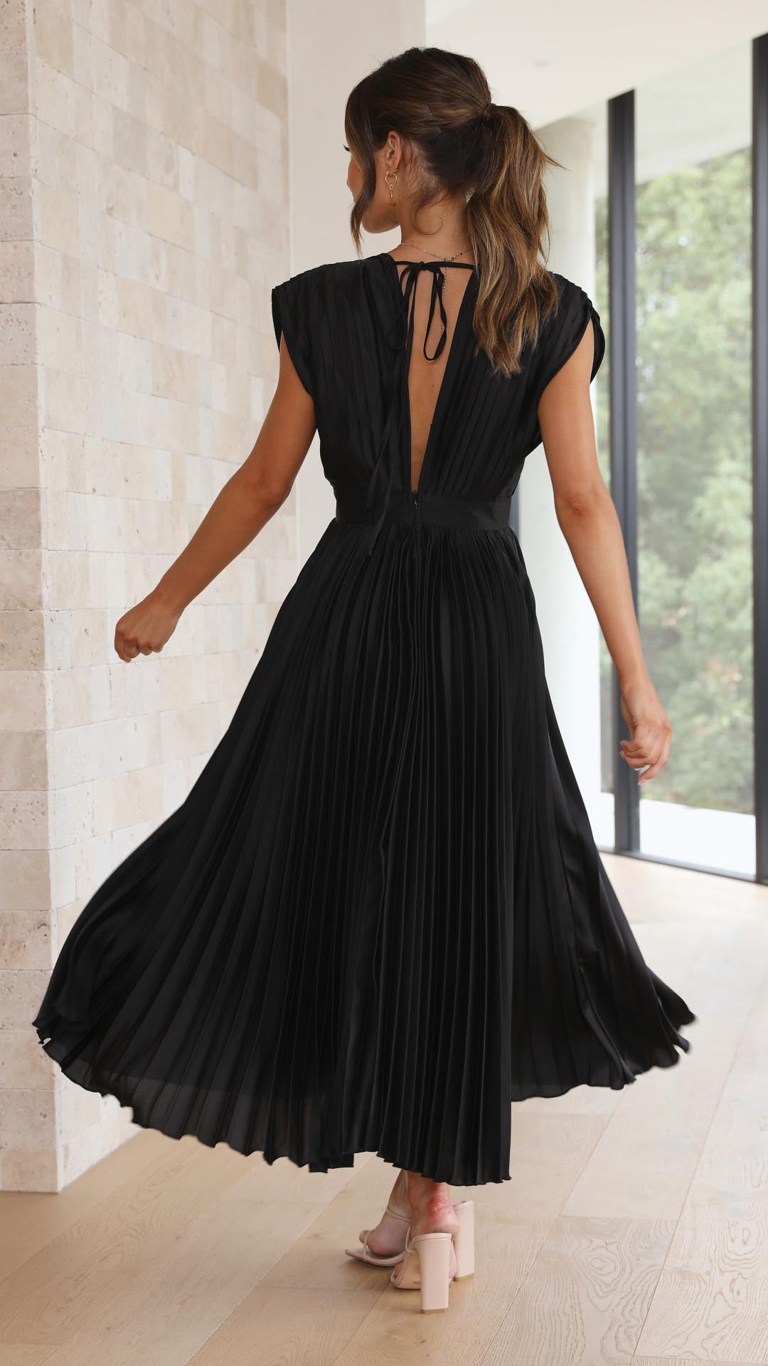 Black - A woman wearing a dusty rose pleated satin midi dress with a deep V-neckline, cap sleeves, and a cinched waist, standing against a textured stone wall.