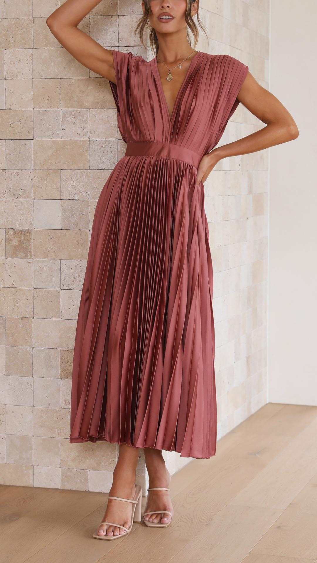 A woman wearing a dusty rose pleated satin midi dress with a deep V-neckline, cap sleeves, and a cinched waist, standing against a textured stone wall.