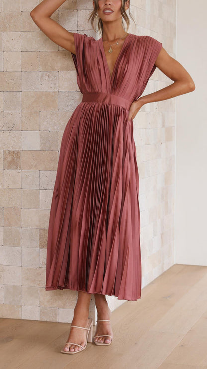 A woman wearing a dusty rose pleated satin midi dress with a deep V-neckline, cap sleeves, and a cinched waist, standing against a textured stone wall.