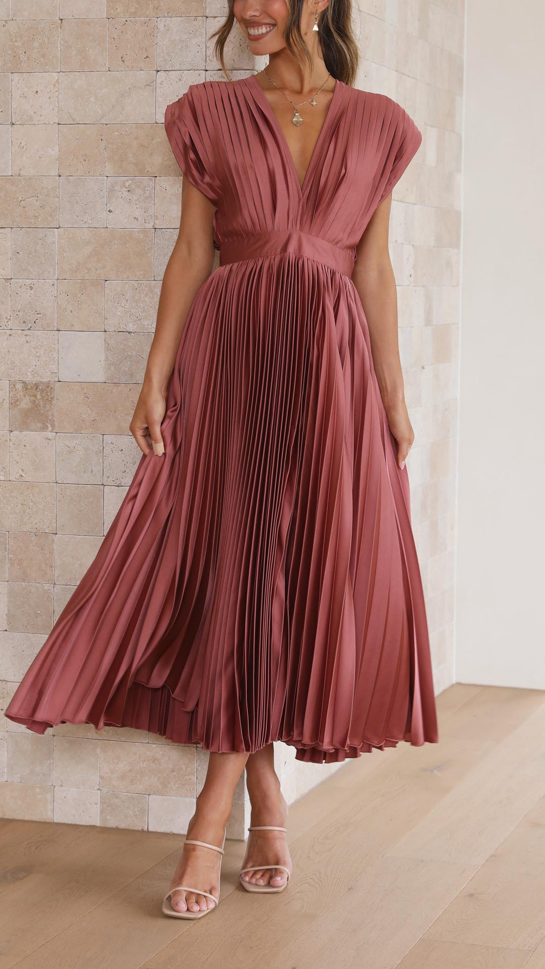 Antique Pink - A woman wearing a dusty rose pleated satin midi dress with a deep V-neckline, cap sleeves, and a cinched waist, standing against a textured stone wall.