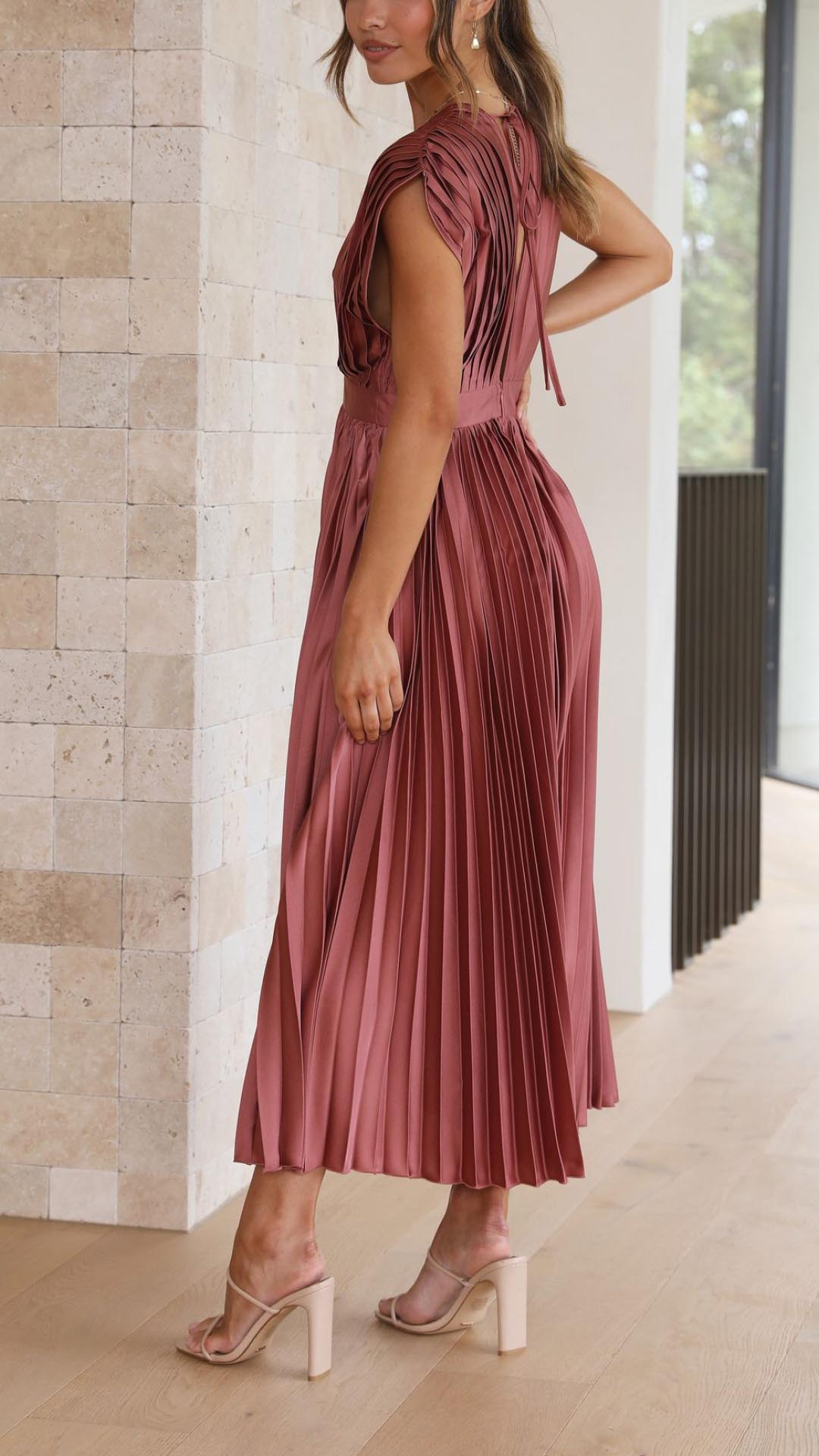 Antique Pink - A woman wearing a dusty rose pleated satin midi dress with a deep V-neckline, cap sleeves, and a cinched waist, standing against a textured stone wall.