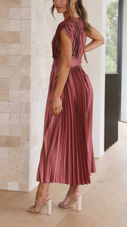 Antique Pink - A woman wearing a dusty rose pleated satin midi dress with a deep V-neckline, cap sleeves, and a cinched waist, standing against a textured stone wall.