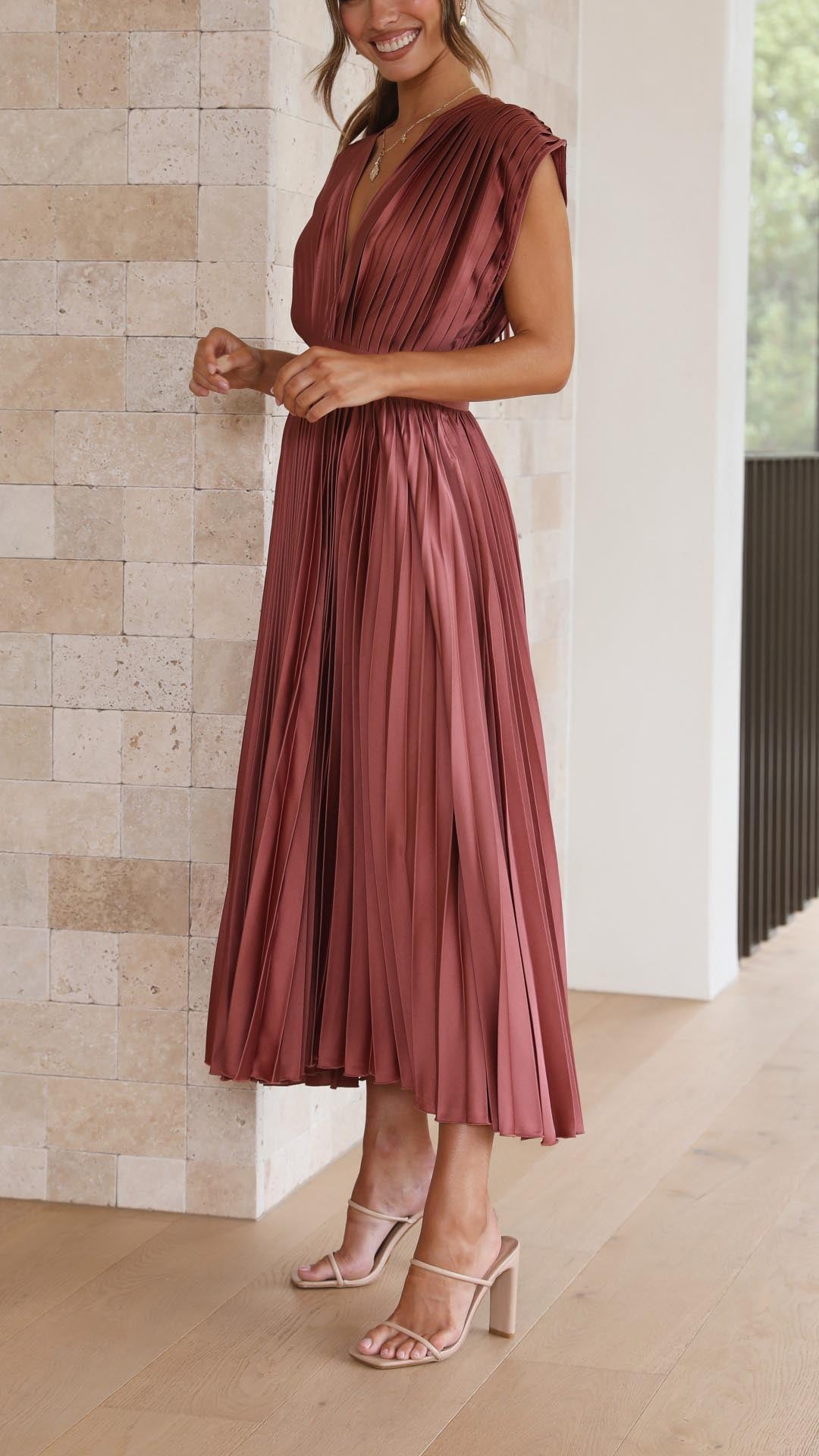 Antique Pink - A woman wearing a dusty rose pleated satin midi dress with a deep V-neckline, cap sleeves, and a cinched waist, standing against a textured stone wall.