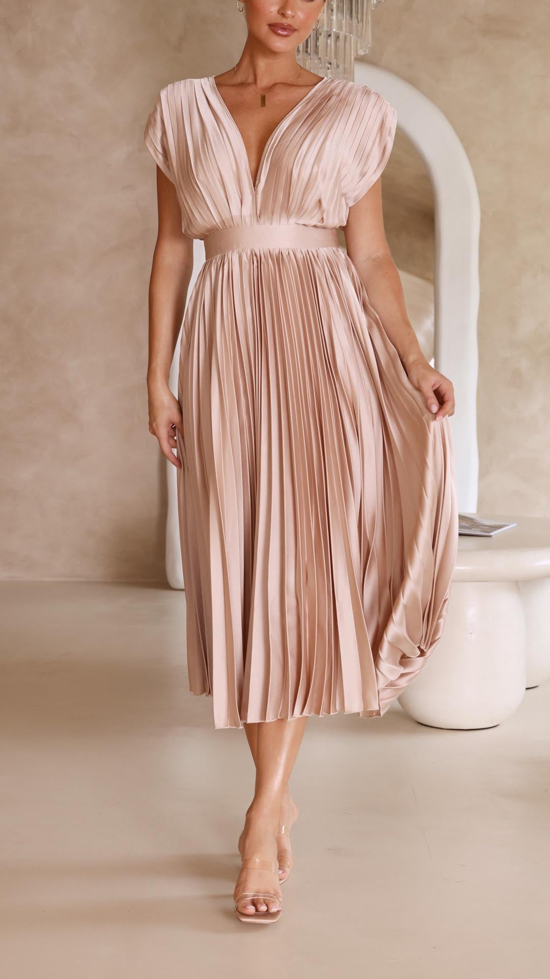 Antique Pink - A woman wearing a dusty rose pleated satin midi dress with a deep V-neckline, cap sleeves, and a cinched waist, standing against a textured stone wall.