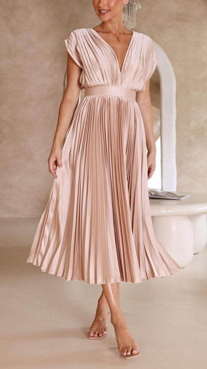 Champagne - A woman wearing a dusty rose pleated satin midi dress with a deep V-neckline, cap sleeves, and a cinched waist, standing against a textured stone wall.