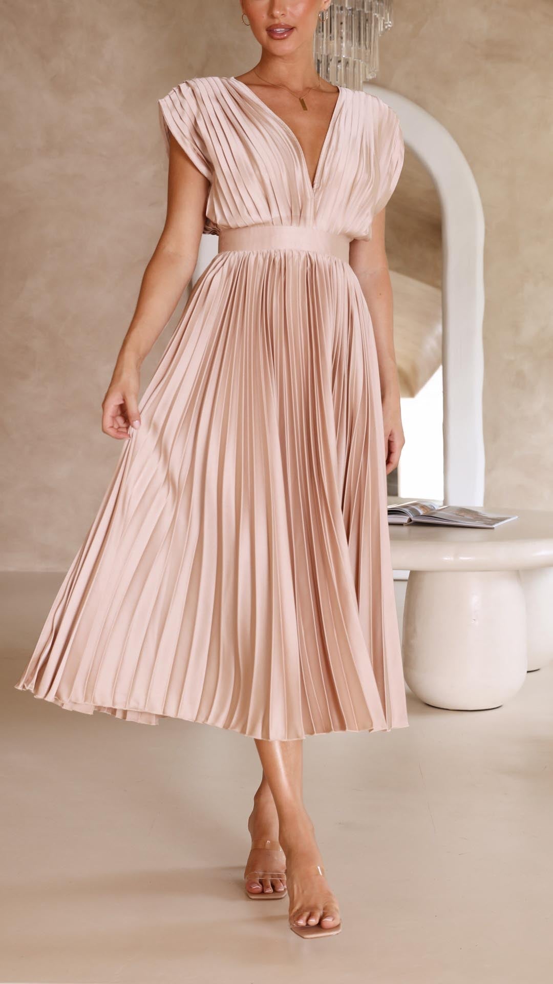 Champagne - A woman wearing a dusty rose pleated satin midi dress with a deep V-neckline, cap sleeves, and a cinched waist, standing against a textured stone wall.