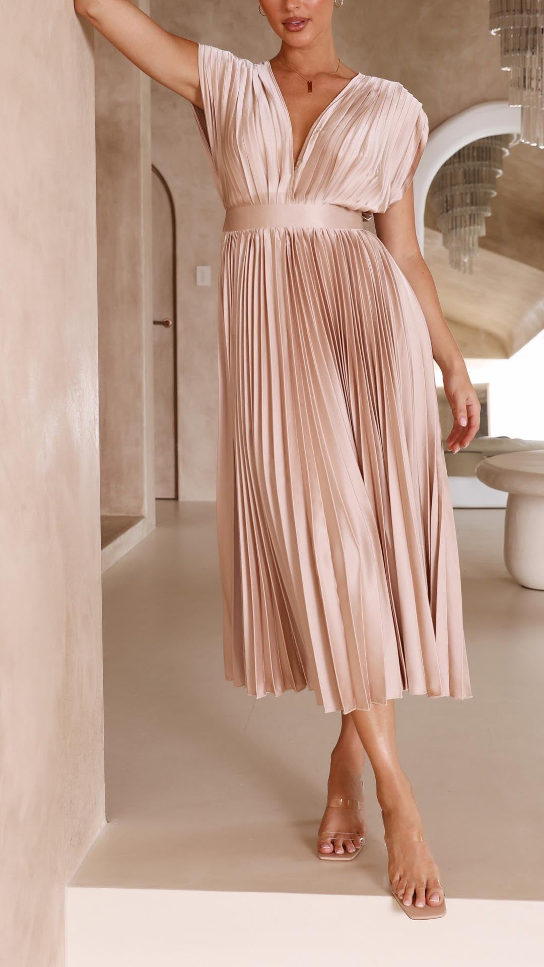 Champagne - A woman wearing a dusty rose pleated satin midi dress with a deep V-neckline, cap sleeves, and a cinched waist, standing against a textured stone wall.