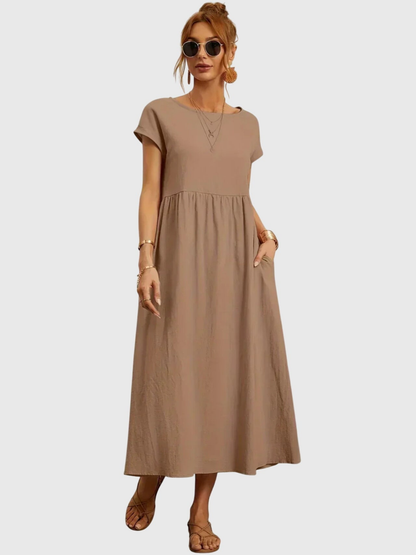 Khaki - Woman wearing a solid short sleeve babydoll maxi dress with a gathered waist and hands resting in side pockets.