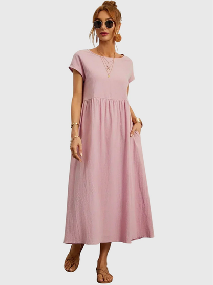 Pink - Woman wearing a solid short sleeve babydoll maxi dress with a gathered waist and hands resting in side pockets.