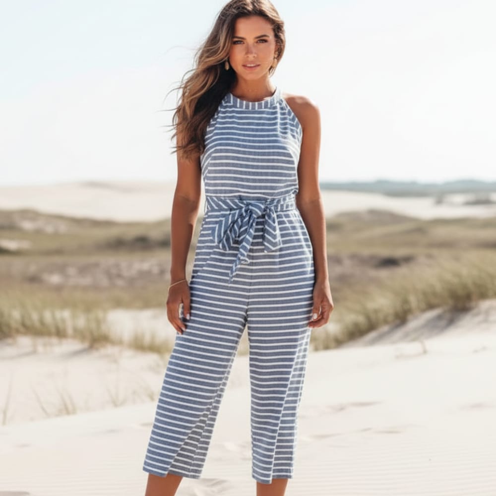 Blue - A woman posing in a desert landscape wearing a grey and white horizontally striped knit jumpsuit featuring a high halter neck, a self-tie waist belt, and cropped wide-leg pants.