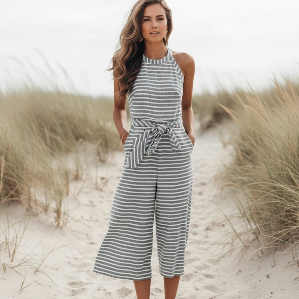 Gray - A woman posing in a desert landscape wearing a grey and white horizontally striped knit jumpsuit featuring a high halter neck, a self-tie waist belt, and cropped wide-leg pants.