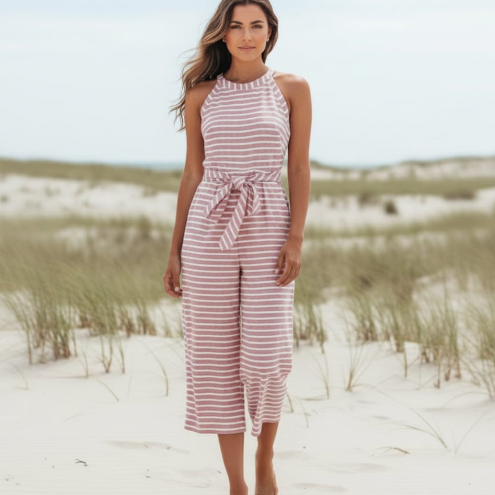 Pink - A woman posing in a desert landscape wearing a grey and white horizontally striped knit jumpsuit featuring a high halter neck, a self-tie waist belt, and cropped wide-leg pants.