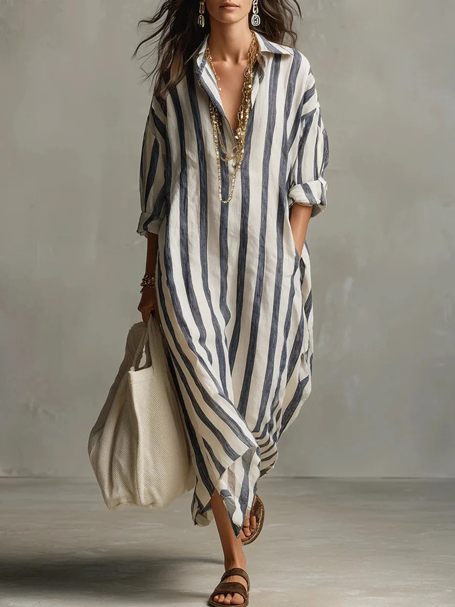Blue - A woman walks wearing an oversized, vertical-striped linen maxi dress in navy and white, featuring a collared V-neck, rolled-up sleeves, and side pockets, paired with a woven tote bag.