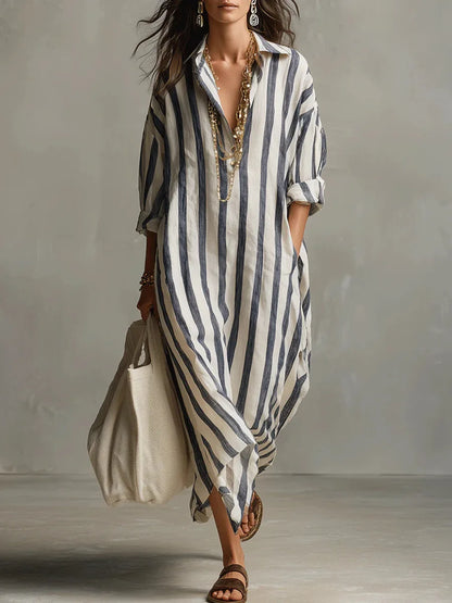 Blue - A woman walks wearing an oversized, vertical-striped linen maxi dress in navy and white, featuring a collared V-neck, rolled-up sleeves, and side pockets, paired with a woven tote bag.