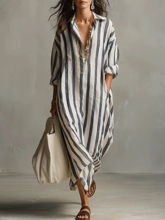 Blue - A woman walks wearing an oversized, vertical-striped linen maxi dress in navy and white, featuring a collared V-neck, rolled-up sleeves, and side pockets, paired with a woven tote bag.