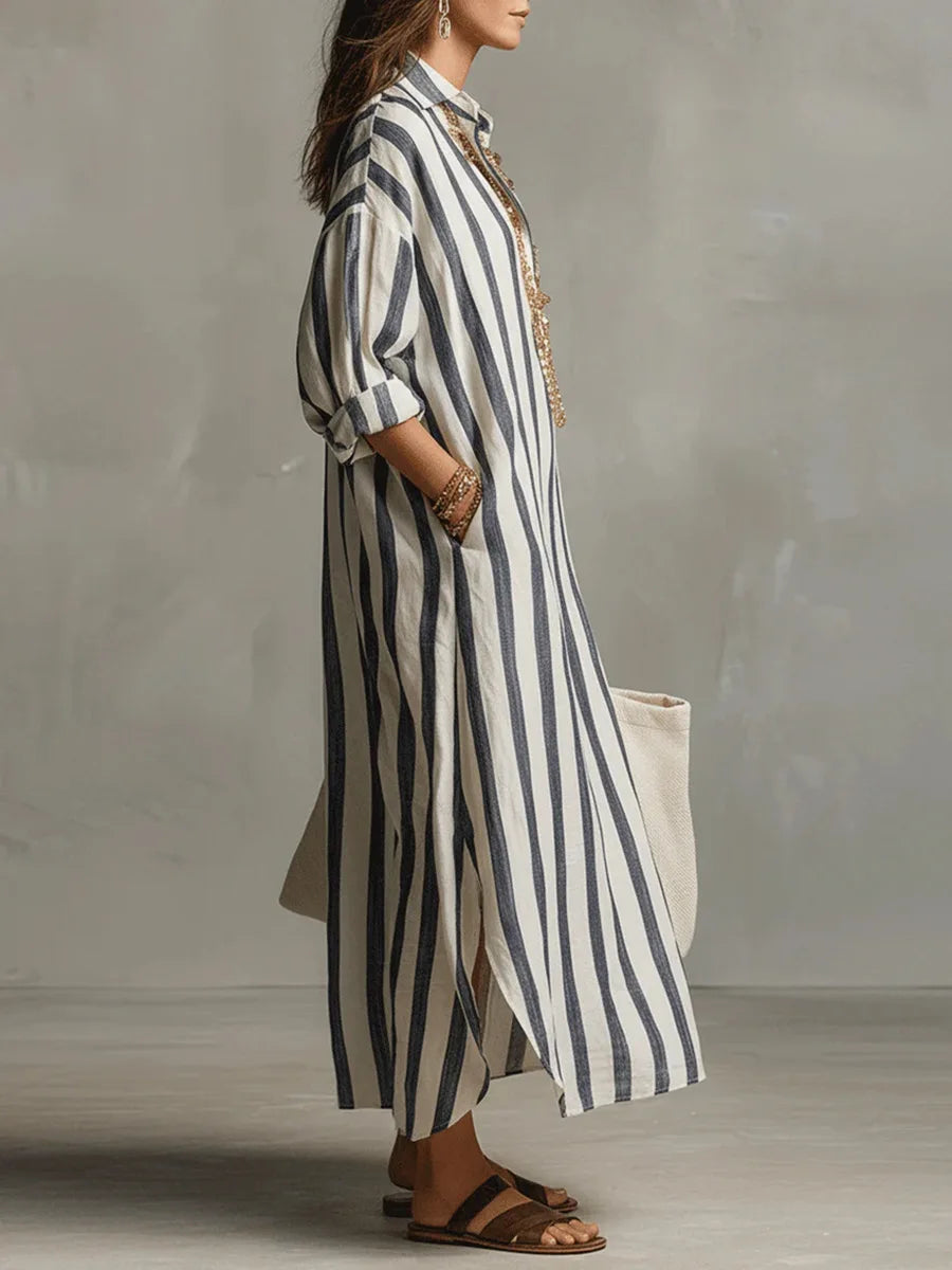 Blue - A woman walks wearing an oversized, vertical-striped linen maxi dress in navy and white, featuring a collared V-neck, rolled-up sleeves, and side pockets, paired with a woven tote bag.