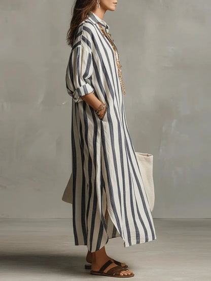 Blue - A woman walks wearing an oversized, vertical-striped linen maxi dress in navy and white, featuring a collared V-neck, rolled-up sleeves, and side pockets, paired with a woven tote bag.