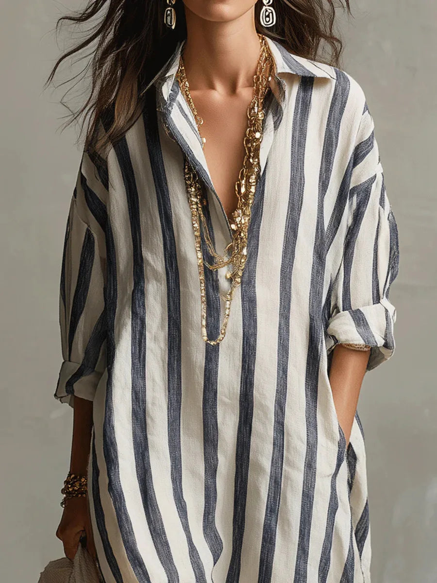 Blue - A woman walks wearing an oversized, vertical-striped linen maxi dress in navy and white, featuring a collared V-neck, rolled-up sleeves, and side pockets, paired with a woven tote bag.