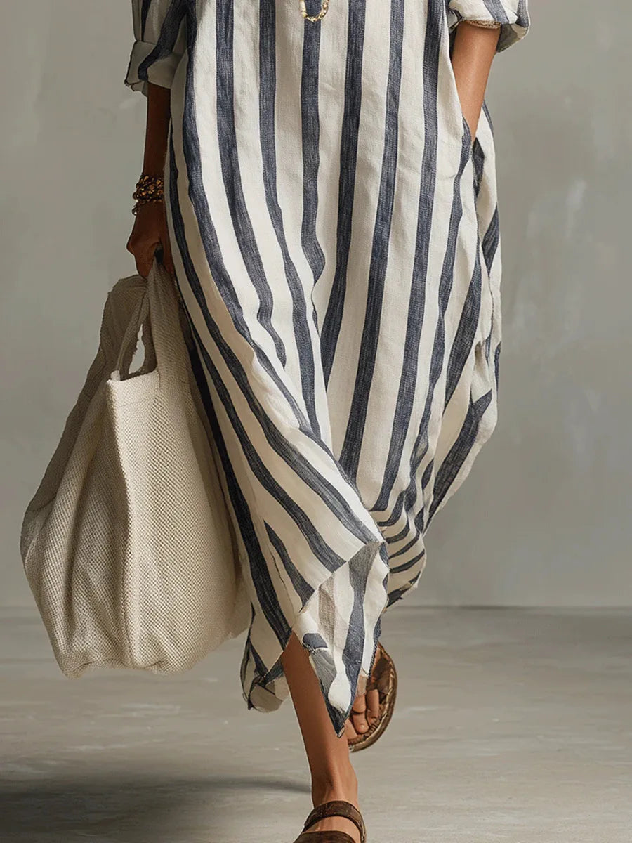 Blue - A woman walks wearing an oversized, vertical-striped linen maxi dress in navy and white, featuring a collared V-neck, rolled-up sleeves, and side pockets, paired with a woven tote bag.