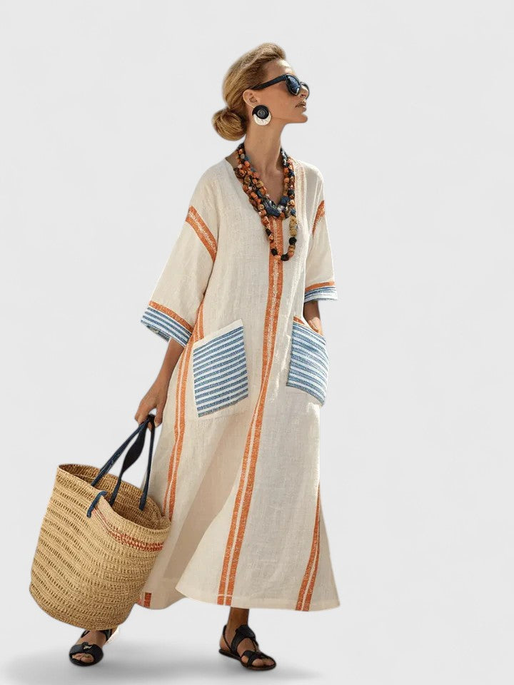 Beige 2 - Woman walking wearing a long, oversized striped maxi dress with half sleeves, front patch pockets, and side slits, carrying a woven tote bag.