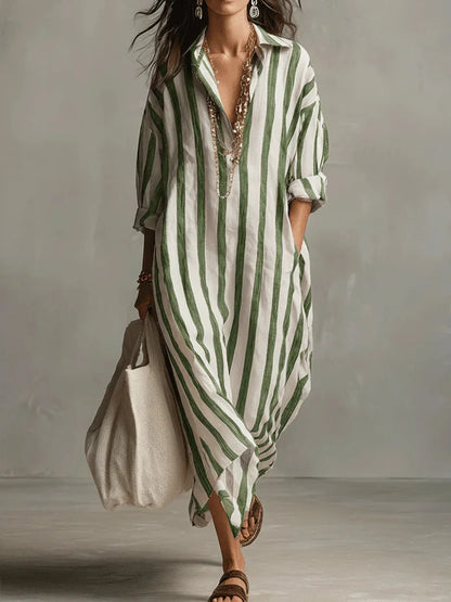 Green - A woman walks wearing an oversized, vertical-striped linen maxi dress in navy and white, featuring a collared V-neck, rolled-up sleeves, and side pockets, paired with a woven tote bag.