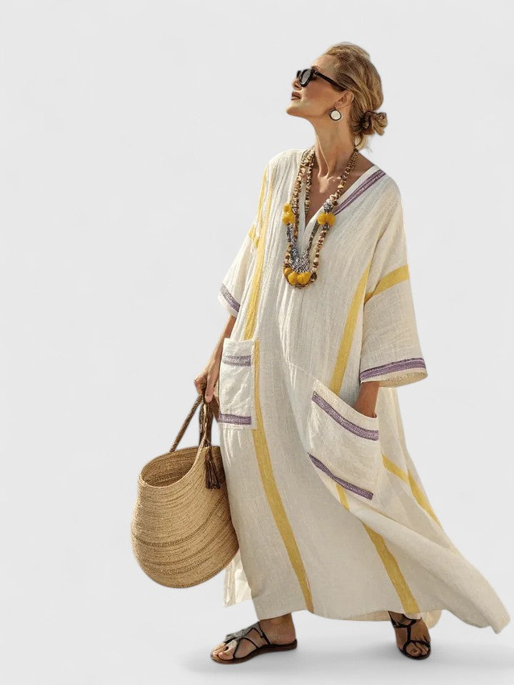 Beige 3 - Woman walking wearing a long, oversized striped maxi dress with half sleeves, front patch pockets, and side slits, carrying a woven tote bag.