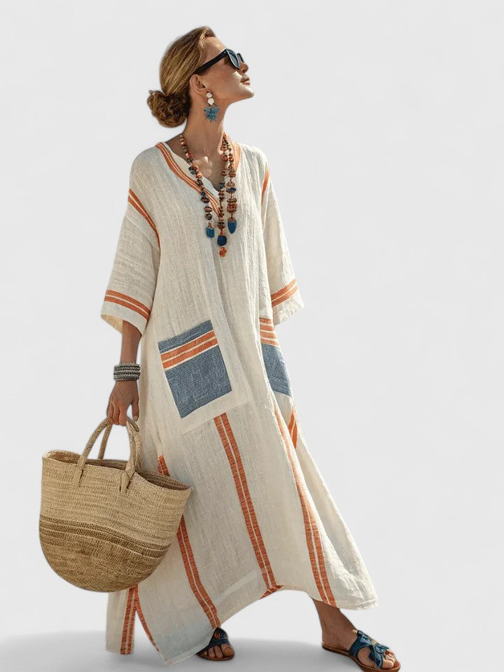 Beige 4 - Woman walking wearing a long, oversized striped maxi dress with half sleeves, front patch pockets, and side slits, carrying a woven tote bag.