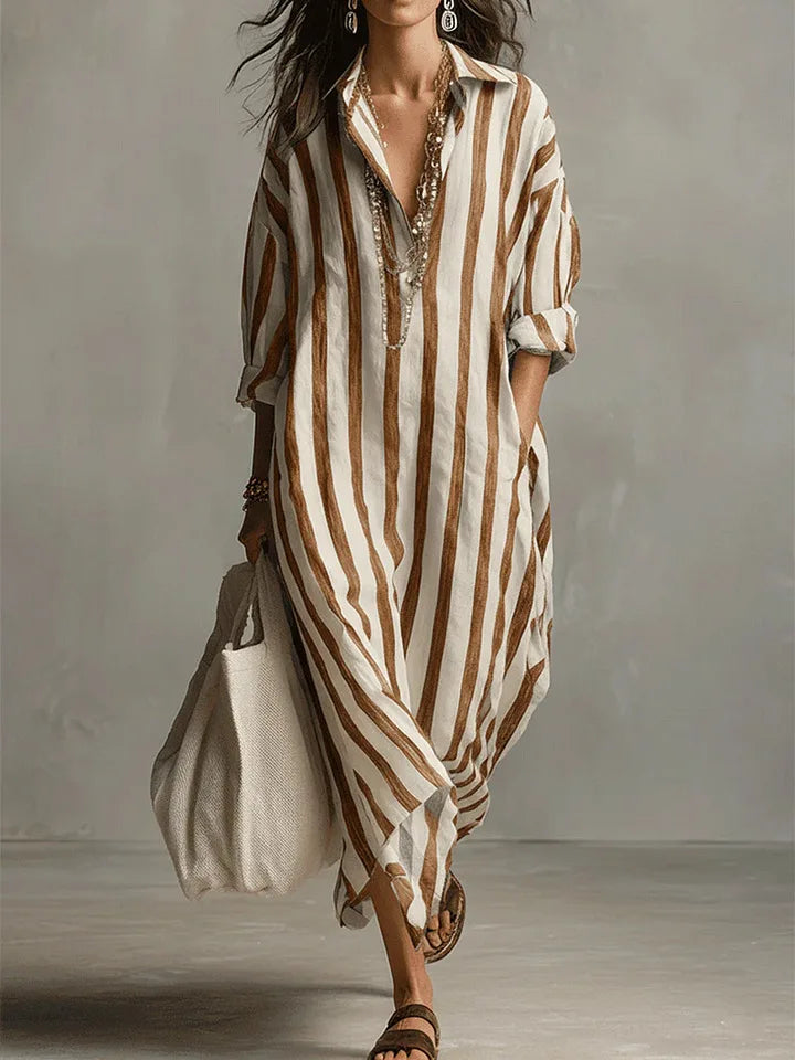 Earthy Yellow - A woman walks wearing an oversized, vertical-striped linen maxi dress in navy and white, featuring a collared V-neck, rolled-up sleeves, and side pockets, paired with a woven tote bag.