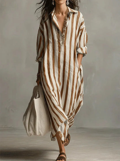 Earthy Yellow - A woman walks wearing an oversized, vertical-striped linen maxi dress in navy and white, featuring a collared V-neck, rolled-up sleeves, and side pockets, paired with a woven tote bag.