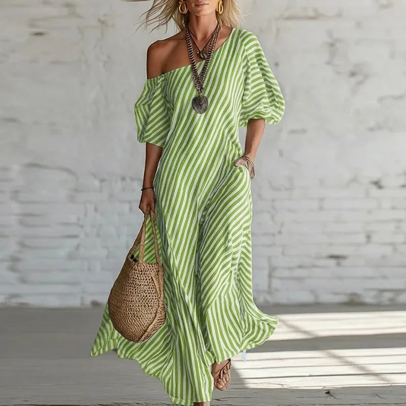 Green - Woman wearing a long, striped, one-shoulder maxi dress with a puff sleeve and a side pocket, holding a woven tote bag.