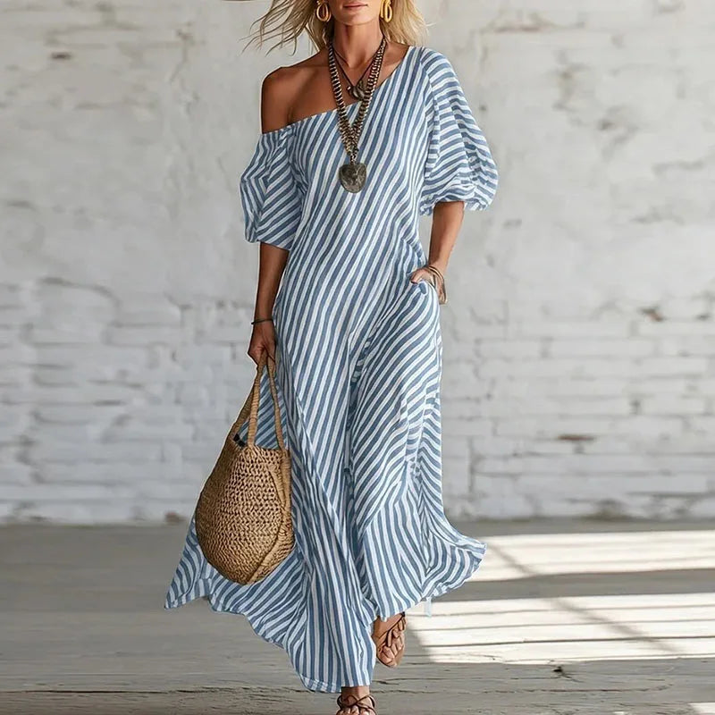 Blue - Woman wearing a long, striped, one-shoulder maxi dress with a puff sleeve and a side pocket, holding a woven tote bag.