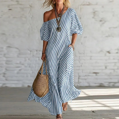 Blue - Woman wearing a long, striped, one-shoulder maxi dress with a puff sleeve and a side pocket, holding a woven tote bag.