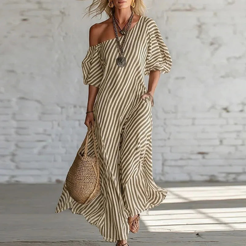 Khaki - Woman wearing a long, striped, one-shoulder maxi dress with a puff sleeve and a side pocket, holding a woven tote bag.
