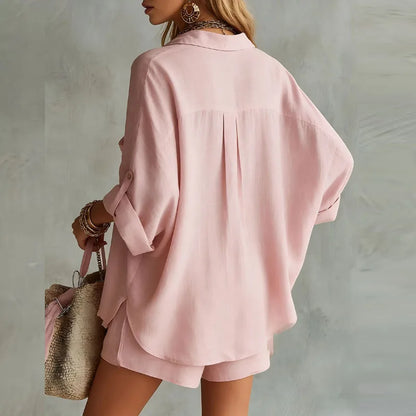 Pink - A woman wearing a dusty pink textured linen-blend two-piece set featuring an oversized long-sleeve button-down tunic shirt with a chest pocket and matching relaxed shorts, accessorized with layered gold necklaces and a woven straw bag.