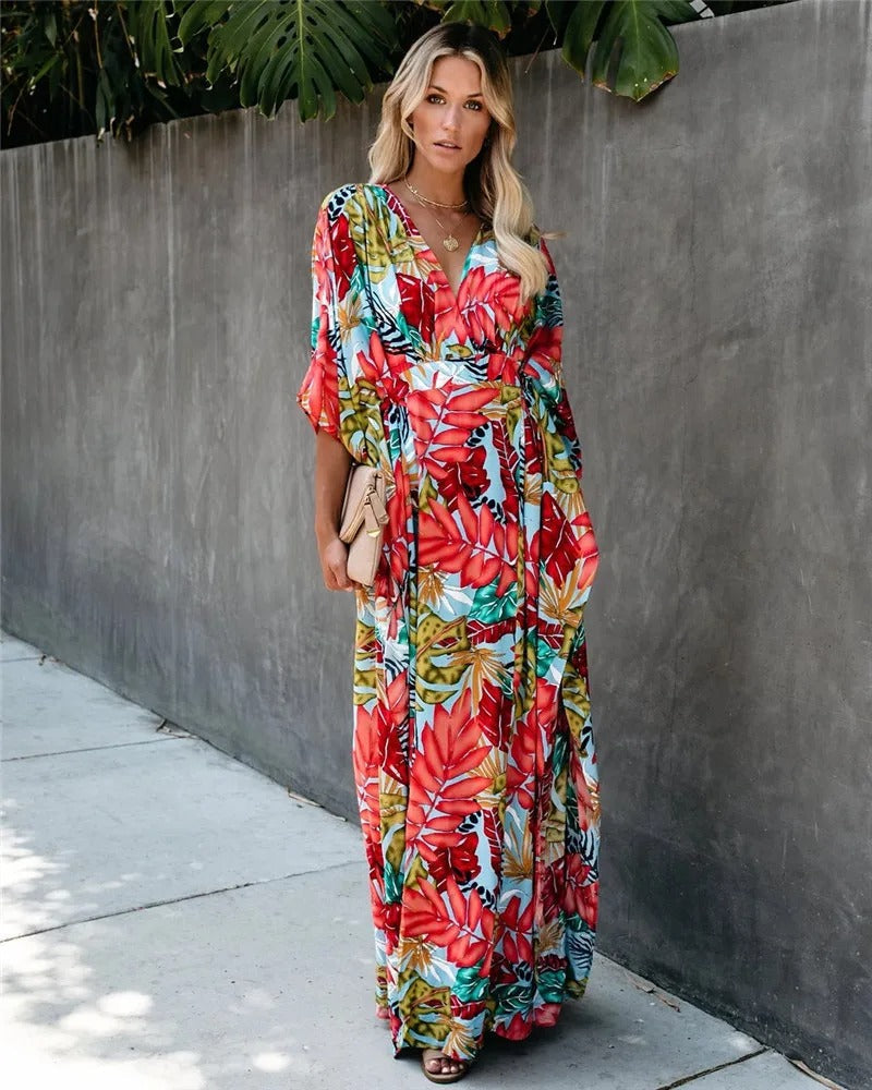 A woman wearing a vibrant tropical botanical print maxi dress with a V-neckline, draped kimono sleeves, and a cinched waist, standing against a grey concrete wall.