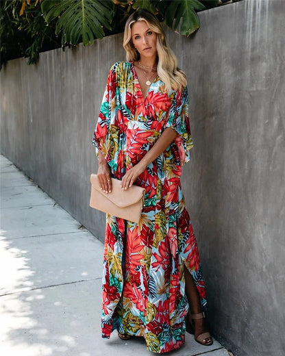 A woman wearing a vibrant tropical botanical print maxi dress with a V-neckline, draped kimono sleeves, and a cinched waist, standing against a grey concrete wall.