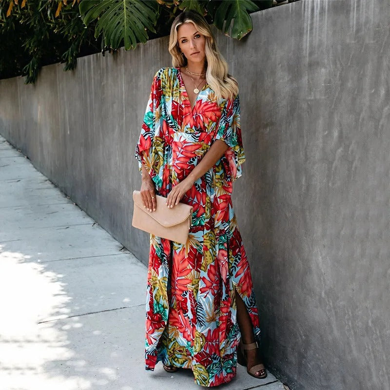A woman wearing a vibrant tropical botanical print maxi dress with a V-neckline, draped kimono sleeves, and a cinched waist, standing against a grey concrete wall.