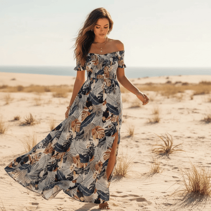 Dark Blue - A woman walking on a sandy beach wearing a tropical leaf print off-the-shoulder maxi dress with a smocked bodice and side slit.