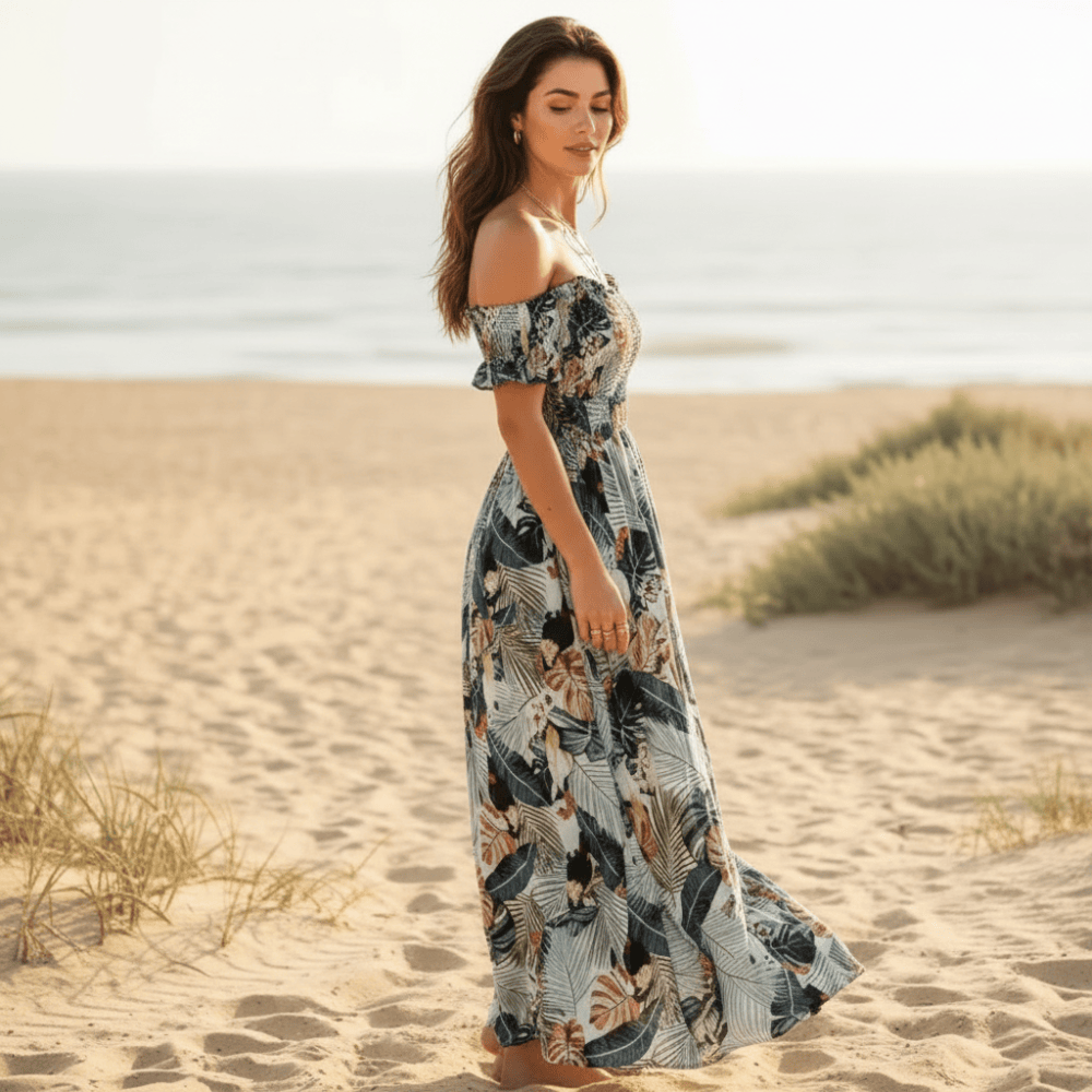 Dark Blue - A woman walking on a sandy beach wearing a tropical leaf print off-the-shoulder maxi dress with a smocked bodice and side slit.