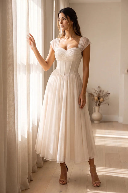 Model wearing a romantic midi dress featuring a ruched sweetheart neckline, sheer cap sleeves, structured corset bodice with visible boning, and a full layered tulle skirt, standing indoors near a sunlit window.