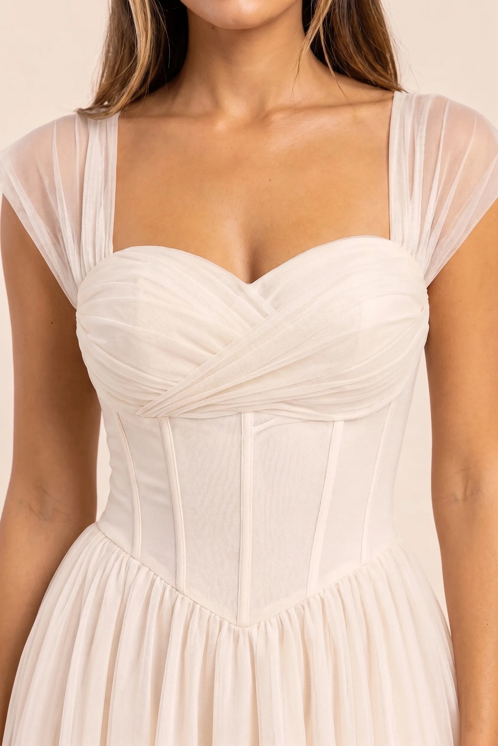 Apricot - Model wearing a romantic midi dress featuring a ruched sweetheart neckline, sheer cap sleeves, structured corset bodice with visible boning, and a full layered tulle skirt, standing indoors near a sunlit window.